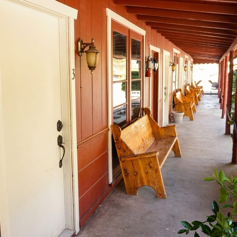 A long, covered outdoor walkway with red wooden walls, white doors, and wooden benches.