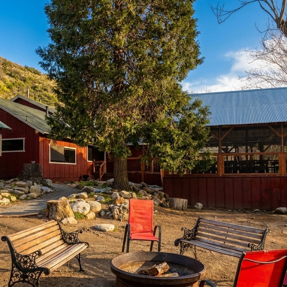 Rustic red buildings with a fire pit and benches in a dirt yard, surrounded by trees and hills.