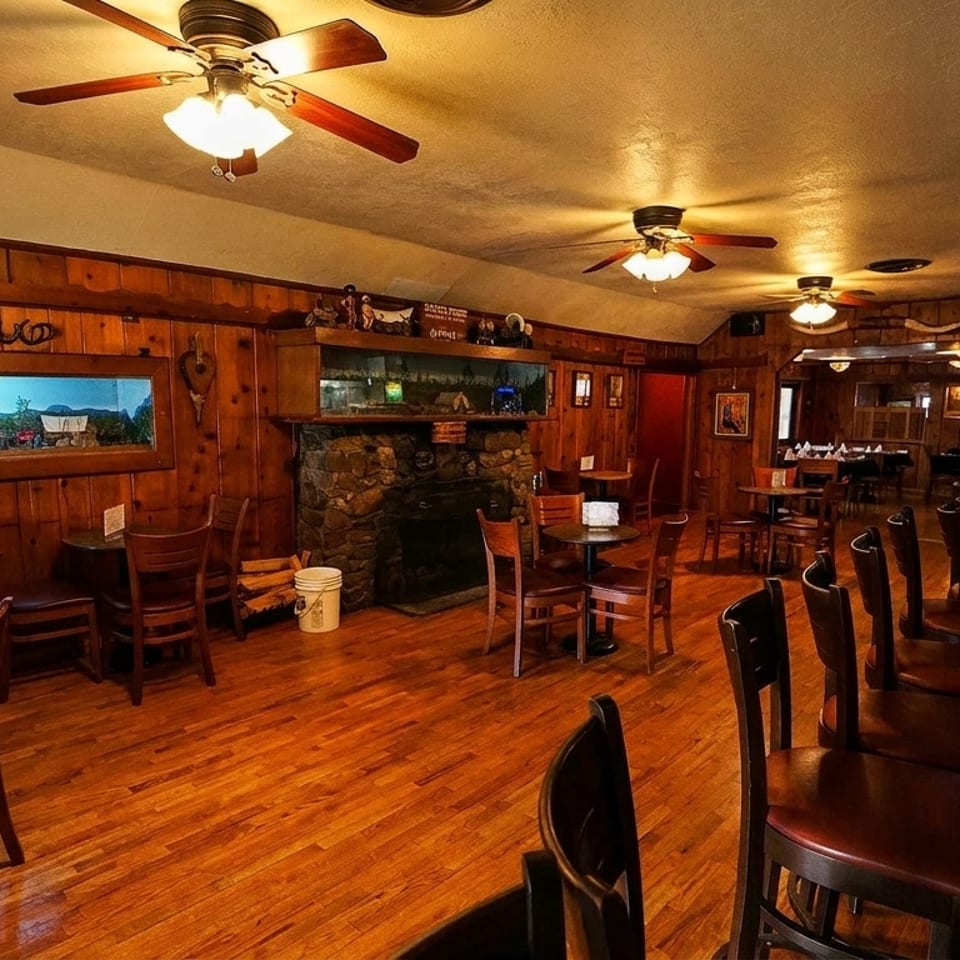 Rustic restaurant interior with wood-paneled walls, a stone fireplace, tables, chairs, and a bar.