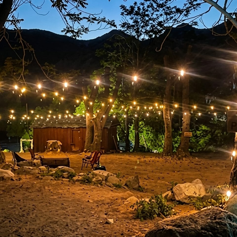 Outdoor dining area at dusk, illuminated by string lights, with trees and mountains in the background.
